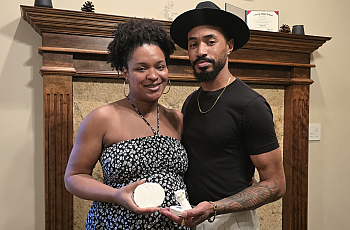 A Black couple stands in front of a wooden mantle holding plaster molds of their late daughters hand and foot.