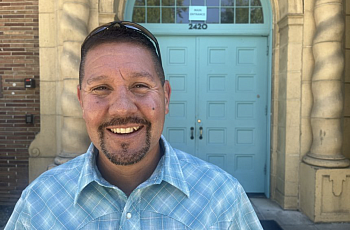 J Vasquez stands in a blue shirt smiling in front of blue double doors.