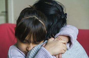 A young girl with bangs hugs an adult tightly, both wearing sweaters, in an emotional moment of comfort and support.