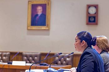 Two women speak into microphones at a formal hearing or meeting, seated at a long table in a government or institutional setting. A portrait and clock are mounted on the wall behind them.