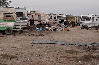 A makeshift encampment of old RVs and trailers surrounded by scattered trash and debris on a dirt lot, indicating homelessness and poverty.