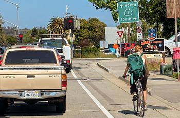 A woman wearing a backpack rides a bicycle in a designated bike lane beside a beige pickup truck at a traffic light intersection. Overhead signs indicate directions for Highway 1 North and South. Traffic is visible in multiple lanes, and there are businesses, including “Tiger Motors,” on the right side of the road. The weather is sunny with clear skies.
