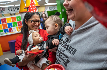 A woman holds a baby with cupcake frosting on their face as another baby leans in; adults around laugh joyfully.