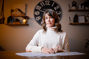 Person in white top with brownish hair in a room sitting on desk with an analog clock begind her