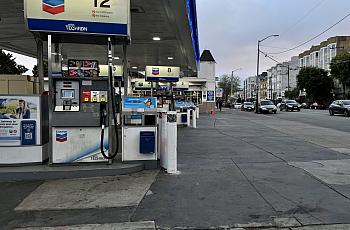A Chevron gas station on a city street at dusk with traffic visible on the adjacent road lined with apartment buildings and trees