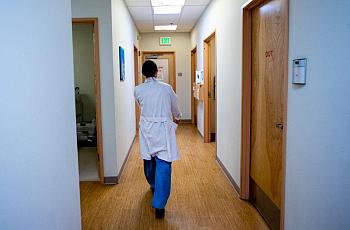 A medical professional in a white coat walks down a brightly lit hallway in a clinic or hospital, with doors labeled “OUT” and an exit sign visible ahead.