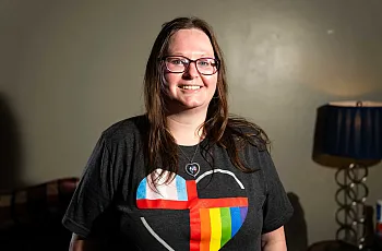 A smiling person with long brown hair and glasses stands indoors wearing a dark T-shirt featuring a heart design