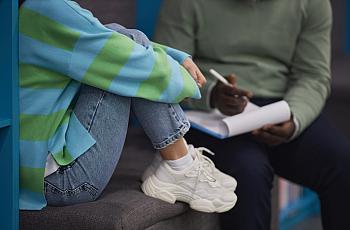 Person sitting with legs close to body, wearing white shoes along with a person writing notes in the background