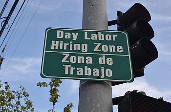 A bilingual street sign reads “Day Labor Hiring Zone / Zona de Trabajo” near a traffic light.
