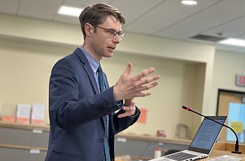 A man in a suit speaks at a podium with a laptop and microphone in a meeting room.