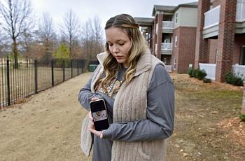 A woman outdoors holds a phone displaying a photo and looks down at it, with apartments and leafless trees in the background.