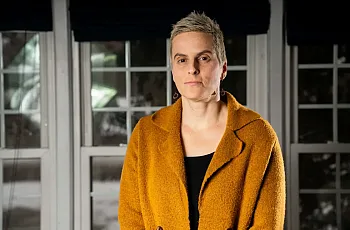 Close-up of a woman in a mustard coat standing indoors with a serious expression, in front of large windows.