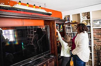 A woman and a child stand in a living room reaching for toys on a shelf beside a TV cabinet, with a large model ship displayed above.
