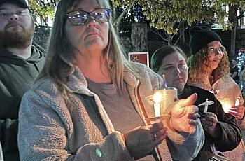 A group of people holds candles at a nighttime vigil; a woman in the foreground wears glasses and a fleece jacket, looking solemnly ahead.
