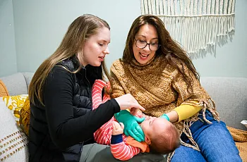 Person holding an infant and another women looking at the infant playfully