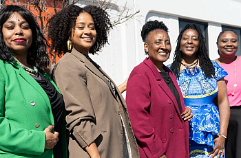  Five women standing outside, smiling in colorful professional outfits.