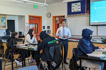 Teacher speaks to students in a science classroom with a projected slide on the board.