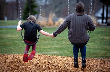 A child and an adult sit on swings holding hands, seen from behind, in a grassy playground with wood chips on the ground.