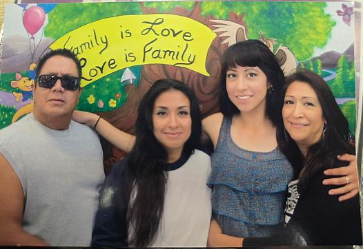 A Latino family of four smiles standing in front of a sign that reads, 'Family is Love, Love is Family'