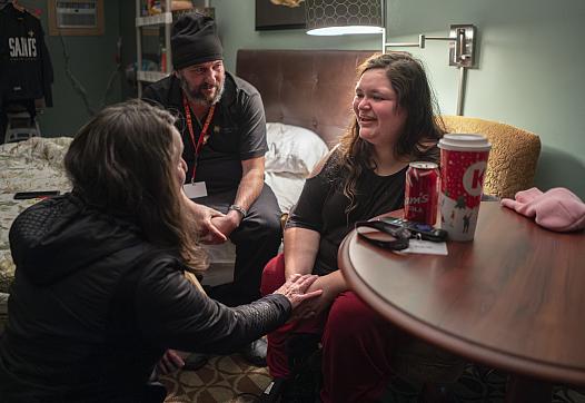 A woman smiles while speaking with two others in a cozy room. One person holds her hands supportively. A soda, coffee cup, and keys rest on the nearby table.