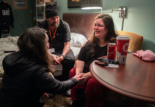 Three people sitting inside a room, one besides a table, one on a bed talking to each other.