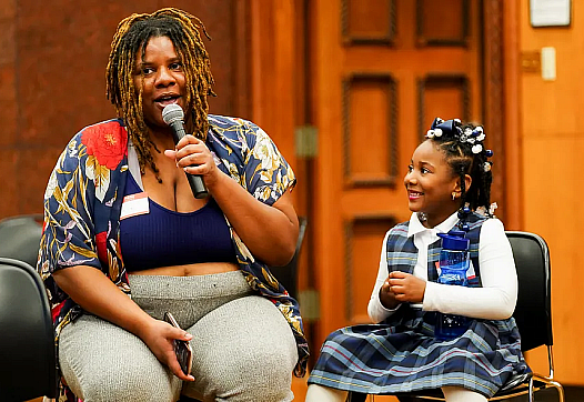 A child seated next to person speaking into a microphone