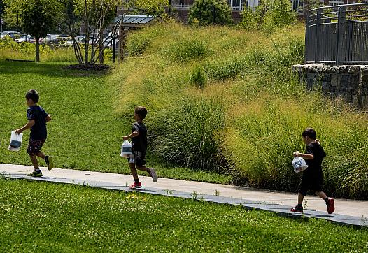 Three children run through a park with bags in their hands.