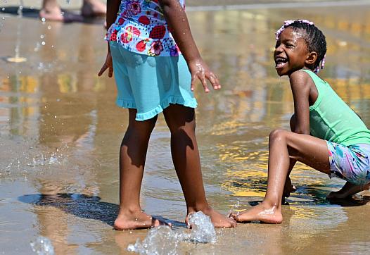 A child plays with a friend in a sprinkler.