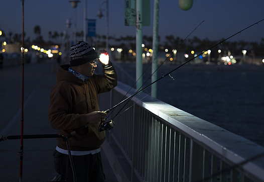 A person wearing a brown hoodie and knit hat is night fishing on a pier, illuminated by a headlamp. They are holding a fishing rod and standing by a railing, with the ocean and city lights visible in the background.