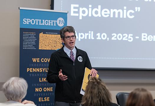 A man in glasses and a black jacket speaks to an audience while holding papers. A presentation screen and event banner are behind him.