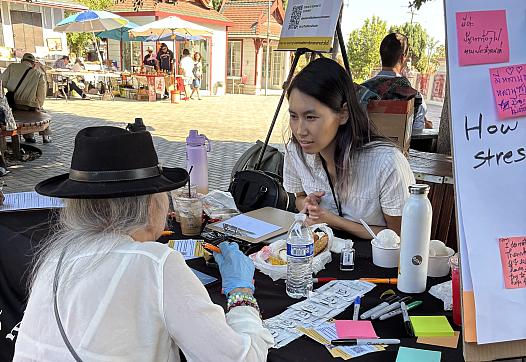 A woman speaks with another person at an outdoor table with pamphlets and notes, next to a mental health sign, at a sunny community market.
