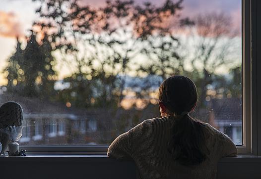 Person staring outside window at a scenery of tress and houses with twilight in the background