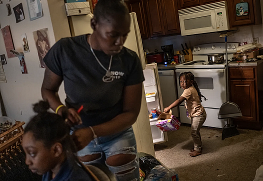 Person doing a child's hair while another child pulls a box out of the refrigerator