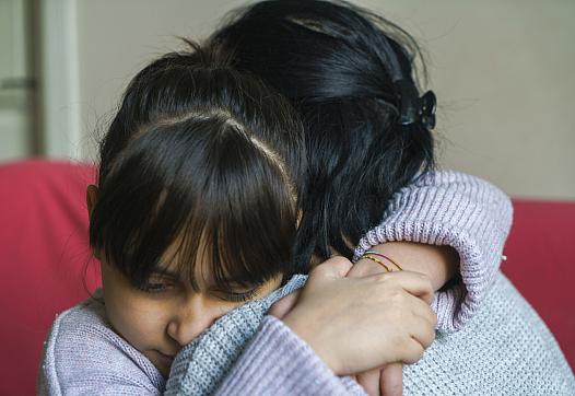 A young girl with bangs hugs an adult tightly, both wearing sweaters, in an emotional moment of comfort and support.