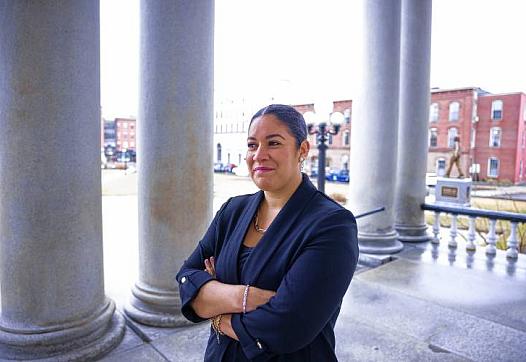 A woman stands confidently with arms crossed on a stone building’s porch, framed by large columns and historic brick buildings in the background.