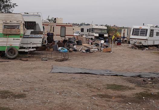 A makeshift encampment of old RVs and trailers surrounded by scattered trash and debris on a dirt lot, indicating homelessness and poverty.