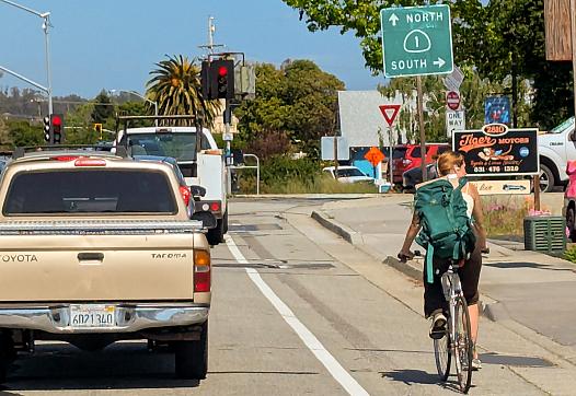 A woman wearing a backpack rides a bicycle in a designated bike lane beside a beige pickup truck at a traffic light intersection. Overhead signs indicate directions for Highway 1 North and South. Traffic is visible in multiple lanes, and there are businesses, including “Tiger Motors,” on the right side of the road. The weather is sunny with clear skies.
