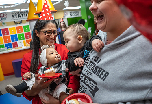 A woman holds a baby with cupcake frosting on their face as another baby leans in; adults around laugh joyfully.