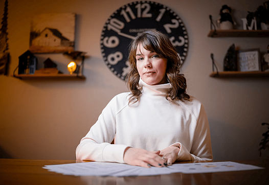 Person in white top with brownish hair in a room sitting on desk with an analog clock begind her