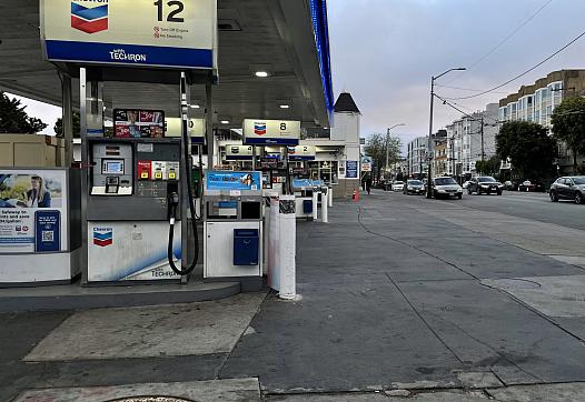 A Chevron gas station on a city street at dusk with traffic visible on the adjacent road lined with apartment buildings and trees