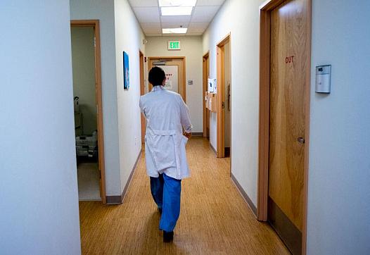 A medical professional in a white coat walks down a brightly lit hallway in a clinic or hospital, with doors labeled “OUT” and an exit sign visible ahead.