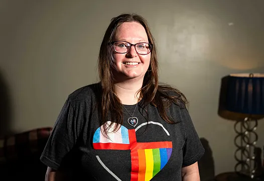 A smiling person with long brown hair and glasses stands indoors wearing a dark T-shirt featuring a heart design