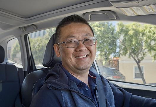 Person sitting in the driving seat of a car with a blue jacket and well cropped hair with spectacles