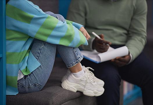 Person sitting with legs close to body, wearing white shoes along with a person writing notes in the background