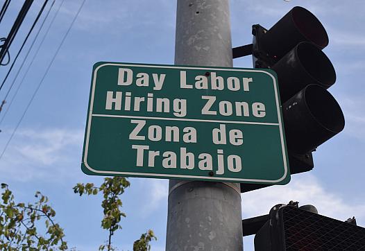 A bilingual street sign reads “Day Labor Hiring Zone / Zona de Trabajo” near a traffic light.