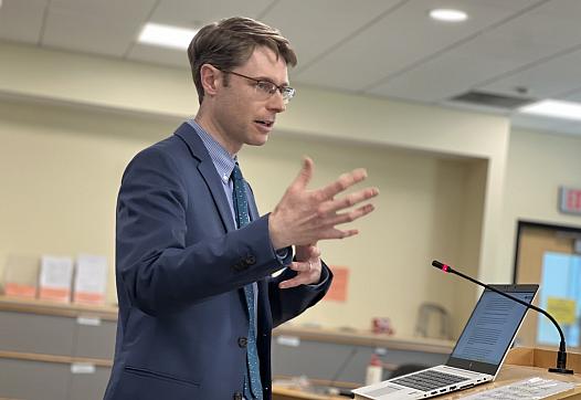 A man in a suit speaks at a podium with a laptop and microphone in a meeting room.