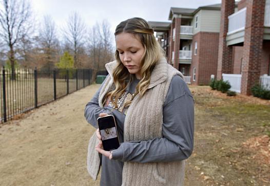 A woman outdoors holds a phone displaying a photo and looks down at it, with apartments and leafless trees in the background.