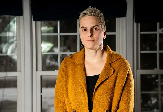 Close-up of a woman in a mustard coat standing indoors with a serious expression, in front of large windows.