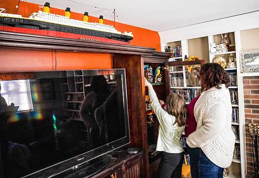 A woman and a child stand in a living room reaching for toys on a shelf beside a TV cabinet, with a large model ship displayed above.