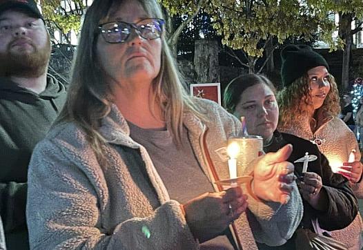 A group of people holds candles at a nighttime vigil; a woman in the foreground wears glasses and a fleece jacket, looking solemnly ahead.